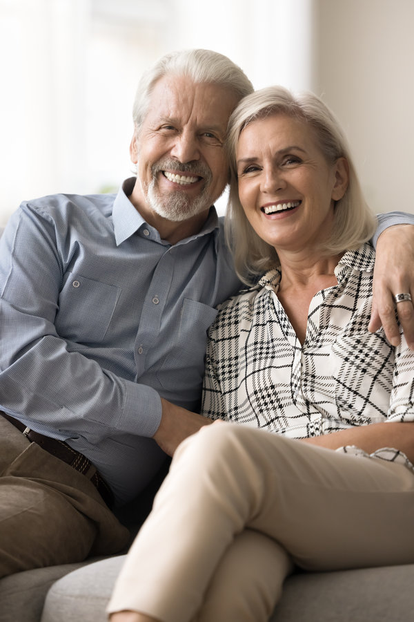 Cheerful positive senior retired husband and wife hugging on home sofa, looking at camera with toothy smiles, showing healthy white teeth, promoting elderly dental service. Vertical portrait shot
